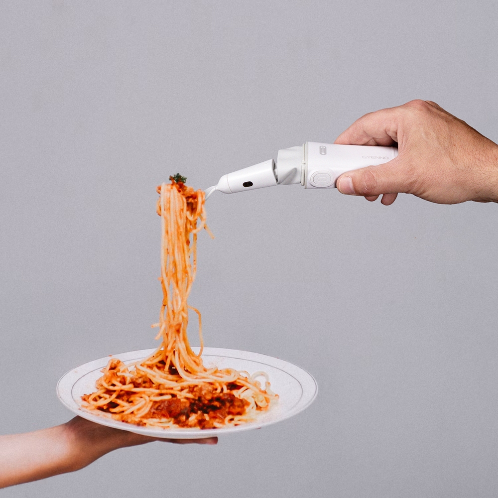Hand holding a white Gyenno utensil with a fork attachment lifting twirled spaghetti above a plate held by another hand 